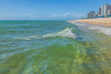 Mavi gökyüzünün altındaki mimari binaların bulunduğu kıyı şeridi manzarasına kumlu sahilde hafif dalgalar eşlik ediyor. Miami Beach, Florida, ABD.
