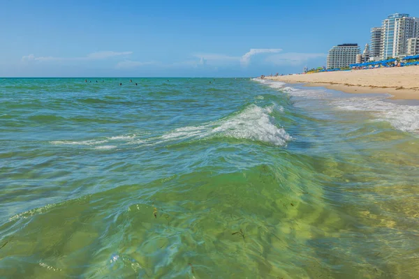 View of shoreline with architectural buildings under blue sky on horizon accompanied by gentle waves rolling onto sandy beach. Miami Beach, Florida, USA.