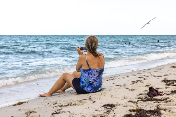 Bikinili bir kadının kumlu sahilde cep telefonuyla pelikan yakaladığı yakın çekim görüntüsü. Miami Plajı. ABD.