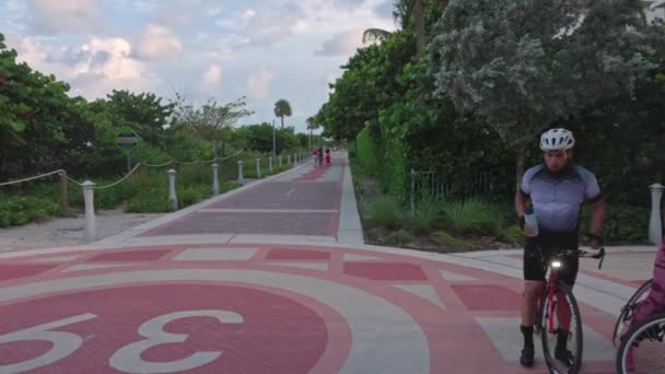 ウォーキングストリートで自転車を運転する家族のカップルの閉鎖的な眺めは 飲料水から飲料水を得ます マイアミビーチ アメリカ合衆国 — ストック動画