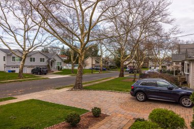 View of a tranquil suburban street with bare trees, family homes, a brick-patterned driveway, and an SUV parked on the side. New Jersey. USA.  clipart