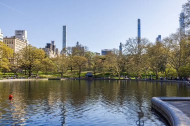 İnsanlar Central Park 'ta göl kenarındaki kaldırımlar boyunca yürüyorlar. Arka planda Manhattan gökdelenleri var. - New York. ABD. 