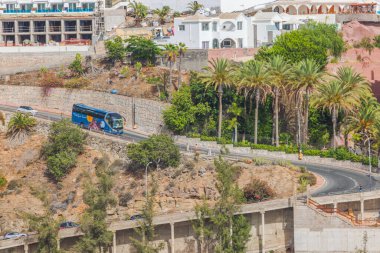 Gran Canaria 'da verimli yeşillikler, palmiye ağaçları ve arka planda konut binalarıyla çevrili bir otobüs ve arabayla güzel bir tepe manzarası. Las Palmas, İspanya. 