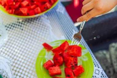 Çocuğun eli, çatalla yakın planda. Masadan dilimlenmiş kırmızı karpuz parçası alıyor.. 