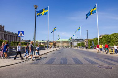 Stockholm 'ün göbeğindeki güzel cadde manzarası İsveç bayrakları, kaldırımlarda yürüyen insanlar ve açık mavi gökyüzünün altındaki ana Kraliyet Sarayı.. 