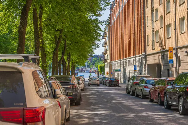 stock image Beautiful view of tree-lined street in Stockholm with parked cars along sides, brick buildings, and glimpse of Norrstrom river in distance on sunny day. 