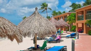 Aruba 'da sazdan şemsiyeler, güneşlikler, palmiye ağaçları ve okyanus kıyısında parçalı bulutlu gökyüzünün altında otel arazisi olan güzel bir sahil manzarası.. 