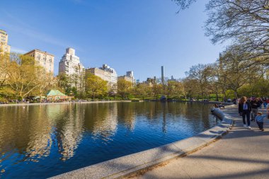 Yayalar, Manhattan 'ın yüksek gökdelenleri tarafından çerçevelenmiş Central Park' taki göl kenarındaki patikalar boyunca yürürler. - New York. ABD.