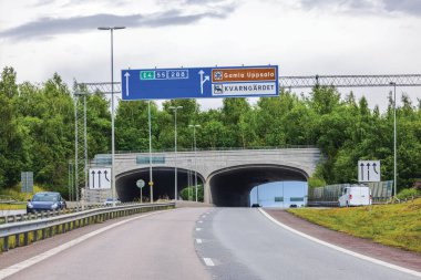 Bulutlu arka planda yeşil ağaçlarla çevrili tünele giden yol işaretleri olan otoyol. İsveç.
