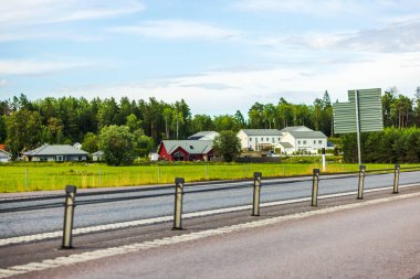 Şeritler arasında çitlerle çevrili, tarım arazilerine ve modern villaları olan köye karşı kurulmuş güzel bir otoyol manzarası. İsveç.