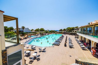Yüzen ve güneşlenen konuklarla dolu bir otel havuzu. Açık mavi gökyüzünün altında güneşlenen şezlonglar, masalar ve sandalyelerle çevrili. Girit. Yunanistan. 