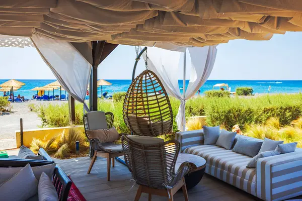 Stock image Outdoor lounge area of hotel with comfortable seating, hanging chair, and curtains, offering view of beach and sea in background. Greece.