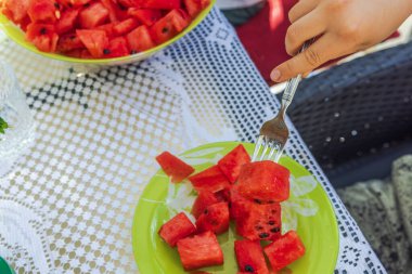 Masadaki bir tabaktan olgun kırmızı karpuzu almak için çatal kullanan bir çocuğun elini yakın plan çek..