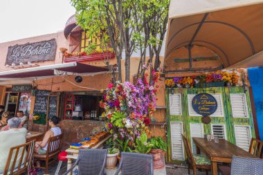 View of outdoor cafe in Curacao with colorful flowers decorating facade, tables with people dining, and vibrant signs. clipart