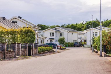 Güneşli bir günde, banliyöde beyaz evler, yeşillikler ve park edilmiş arabaların olduğu sokak manzarası. İsveç, Avrupa.