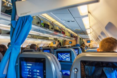 View of airplane economy class cabin, showing passengers seated comfortably while onboard entertainment screens are visible on seat backs.  clipart