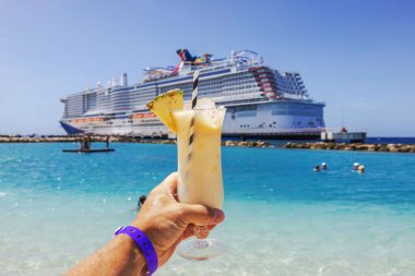 Close-up view of hand holding glass of alcoholic cocktail on Caribbean beach with cruise ship in background. clipart