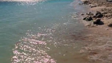 Karayip Denizi 'nden gelen hafif dalgalar Aruba adasındaki parlak güneş ışığı yansımalarıyla kumlu ve kayalık bir plaja vuruyor..