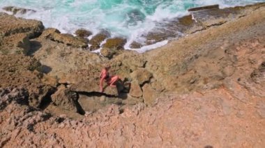 Mayo giymiş bir kız güneşli bir günde Aruba adasında Karayip Denizi kıyısında keskin kayalıklarda dikkatlice yürüyor.. 