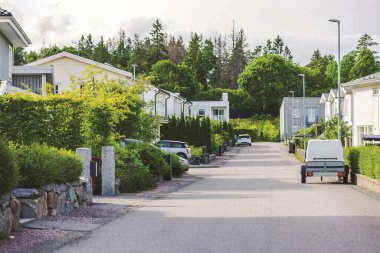 Bulutlu bir günde modern evler, yeşil çitler ve park edilmiş arabalarla dolu sessiz bir banliyö caddesi. İsveç.