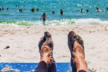 Close up view of feet in sand lying on beach lounge chair with view of people swimming in Atlantic Ocean in Miami Beach. USA. clipart