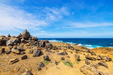 Person walking through rocky landscape along Arubas rugged coast, piles of stones stacked on large boulders near the blue Caribbean Sea. clipart