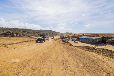 Dirt road with off-road vehicles driving through rugged desert landscape of Aruba, near small huts. clipart