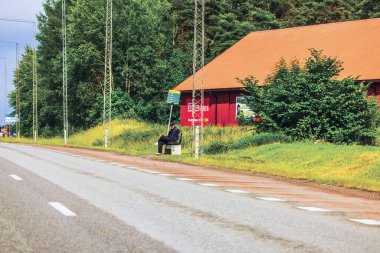 Kırsal otobüs durağında tek başına oturan kişi, kırmızı binanın yanında, yeşil ağaçlar ve çimlerle çevrili, boş yol mesafeye uzanıyor.. 
