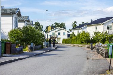 Banliyö caddesinde modern beyaz evler boyunca yürüyen iki kişi, yemyeşil ve açık gökyüzü. İsveç.