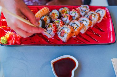 Farklı suşi ruloları ve kalp şeklinde soya sosu tabağıyla renkli tabaktan suşi toplamak için yemek çubukları kullanıyor.. 