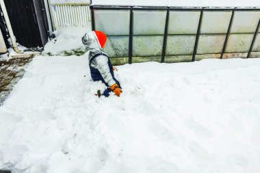 Child wearing winter snowsuit sitting in deep snow near greenhouse in backyard villa. Sweden. clipart