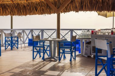 Outdoor beachside restaurant with blue chairs and tables overlooking Caribbean sea, under thatched roof. Curacao.