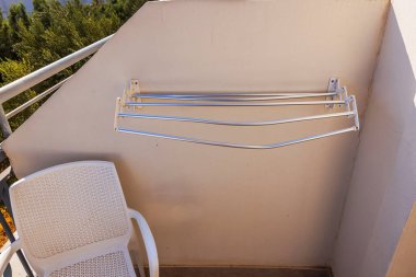 Empty wall-mounted drying rack on sunny balcony with white plastic chair and view of greenery. Crete. Greece. clipart