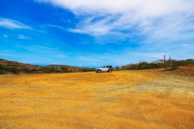 White Jeep on barren desert landscape under wide blue sky in Arikok National Park, Aruba. clipart