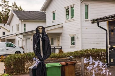 Skeleton Halloween decoration cloaked in black near residential bins, surrounded by faux cobwebs. Sweden. clipart