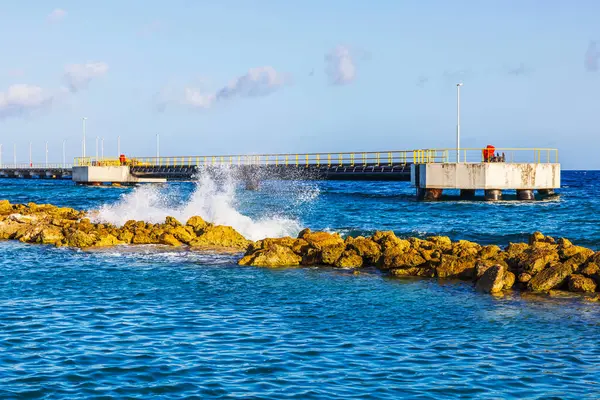 Deniz suyu, dalgakıran rıhtımının yakınındaki kayalara çarpar. Karayipler kıyısındaki berrak mavi gökyüzünün altında. Curacao.