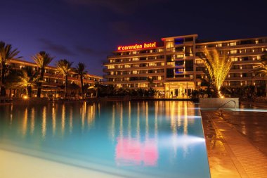 Night view of Corendon Hotel in Curacao with glowing lights, palm trees, and pool reflections creating serene atmosphere.  clipart