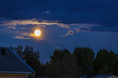 Full moon illuminating dark night sky with soft clouds and silhouettes of trees and rooftops. Sweden. clipart