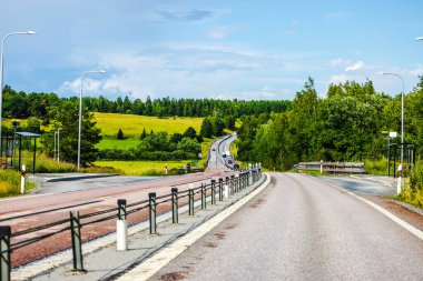 Bulutlu arka planda yeşil tarlaları, ağaçları ve uzak ormanı olan güzel bir otoyol manzarası. İsveç.