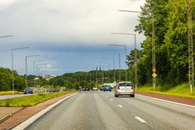 Çevresi yeşil ağaçlarla çevrili otoyol ve bulutlu gökyüzünün altında uzak şehir manzarası. 