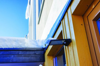 Hareket sensörlü LED aydınlatma donanımı açık mavi gökyüzünün altında, karla kaplı, sarı ahşap duvarlı villanın üzerine monte edilmiş. İsveç.