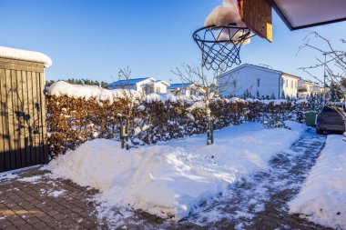 Snow-covered courtyard with cleared path, bare trees, and basketball hoop, surrounded by sunlight and frosty landscape. Sweden. clipart