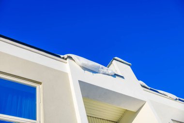 Güneşli kış gününde açık mavi gökyüzünün altında beyaz evin çatısından sarkan kar. İsveç.