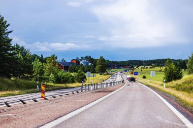 Gökyüzü bulutlu arka planda yeşil tarlalar, ağaçlar ve evler ile çevrili açık kırsal yol. İsveç.