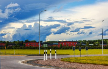 Yeşil tarlalarla ve dramatik bulutlu gökyüzünün altında kırmızı sanayi binalarıyla çevrili yol işaretleriyle dolu bir kavşak. İsveç.
