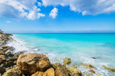 Turkuaz dalgaların açık mavi gökyüzü ve dağınık beyaz bulutlar altında kayalara çarptığı Rocky sahili sahili. Aruba.