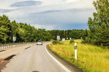 Bulutlu gökyüzünün altındaki yeşil ağaçlar ve tarlalarla çevrili otoban ve asfalt yol boyunca giden araçlar. İsveç.