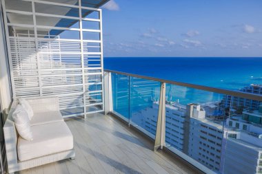 Açık mavi gökyüzünün altında turkuaz Karayip denizine bakan beyaz kanepe ve cam korkuluklu modern otel balkonu. Cancun. Meksika.