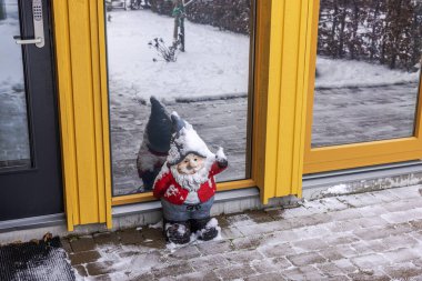 Snow-covered garden gnome standing by entrance door of villa with panoramic reflective windows during winter. Sweden. clipart