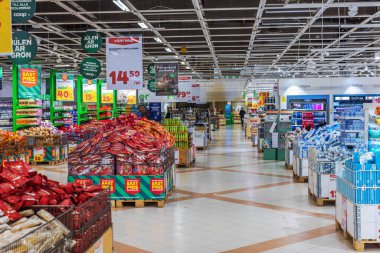 Yığınlanmış ürünler, tanıtım tabelaları, renkli görüntüler ve parlak ışık altında organize edilmiş raflarla süpermarket koridoru. 